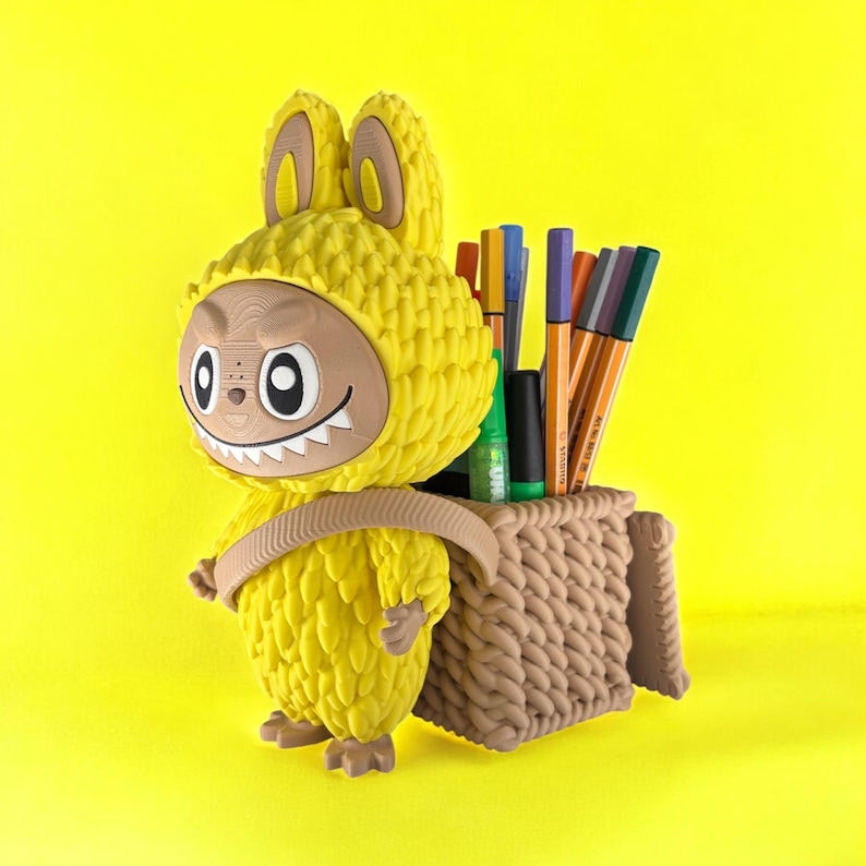 May include: A whimsical yellow pen holder shaped like a creature with a bunny-like hood and a basket on its back. The pen holder is filled with colorful pens and markers, set against a bright yellow background.