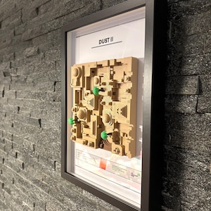 May include: A framed 3D model of the Dust II map from the video game Counter-Strike: Global Offensive. The model is made of beige plastic and features green trees. The frame is black and has a white background with the text "DUST II" printed in black.