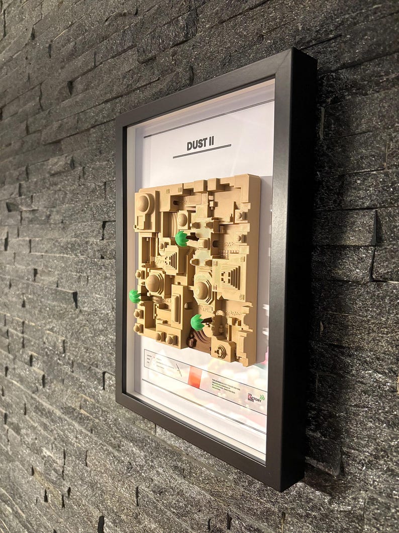 May include: A framed 3D model of the Dust II map from the video game Counter-Strike: Global Offensive. The model is made of beige plastic and features green trees. The frame is black and has a white background with the text "DUST II" printed in black.