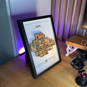 May include: A framed 3D map of a video game level, labeled "CACHE". The map is beige with red and blue accents, set against a white background within a black frame. The frame is displayed on a wooden desk.