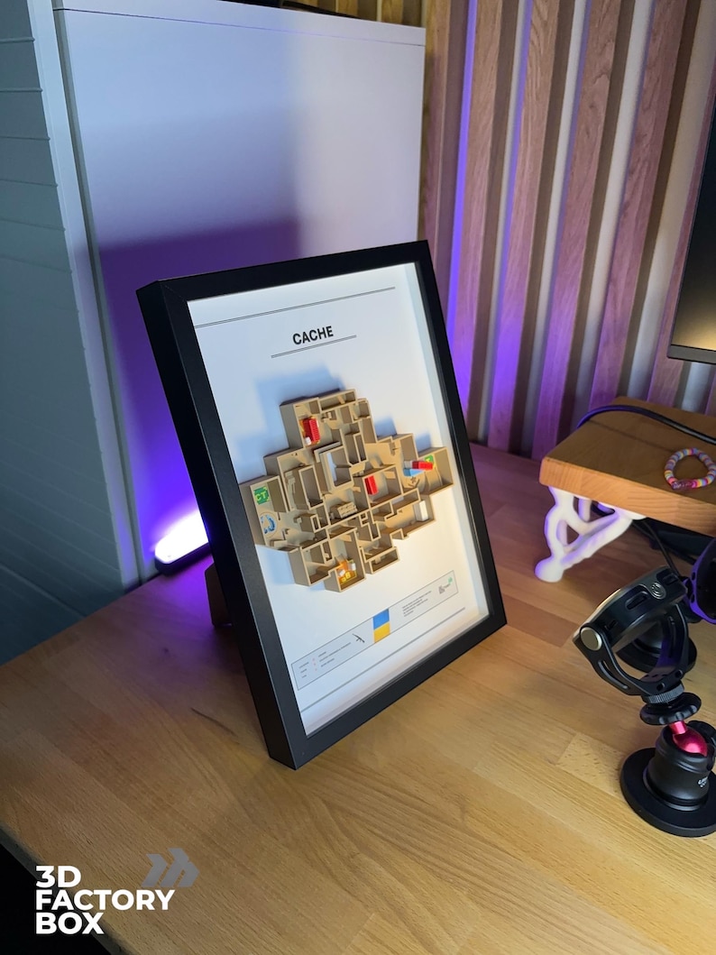 May include: A framed 3D map of a video game level, labeled "CACHE". The map is beige with red and blue accents, set against a white background within a black frame. The frame is displayed on a wooden desk.