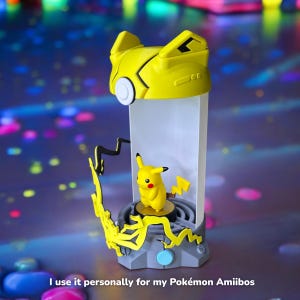 May include: A yellow and gray Pokemon figure display stand with a clear plastic case. The stand features a Pikachu figure inside the case. The stand has a lightning bolt design and a blue button.