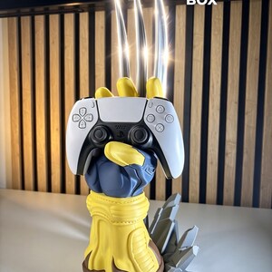 May include: A yellow and blue glove-shaped controller stand featuring three long, metallic silver claws extending upward. The stand holds a white and black wireless game controller. The base is sculpted with gray rock-like details.