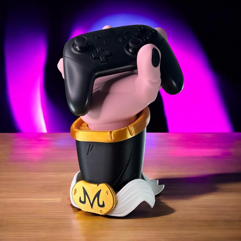 May include: A black video game controller held by a pink hand emerging from a black and gold base. The base has a gold emblem with a stylized "M" and white accents. The background is a gradient of purple and black.