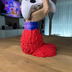 May include: A 3D printed hand holding a white video game controller. The hand is red and has a purple wristband. The hand is standing on a wooden surface.