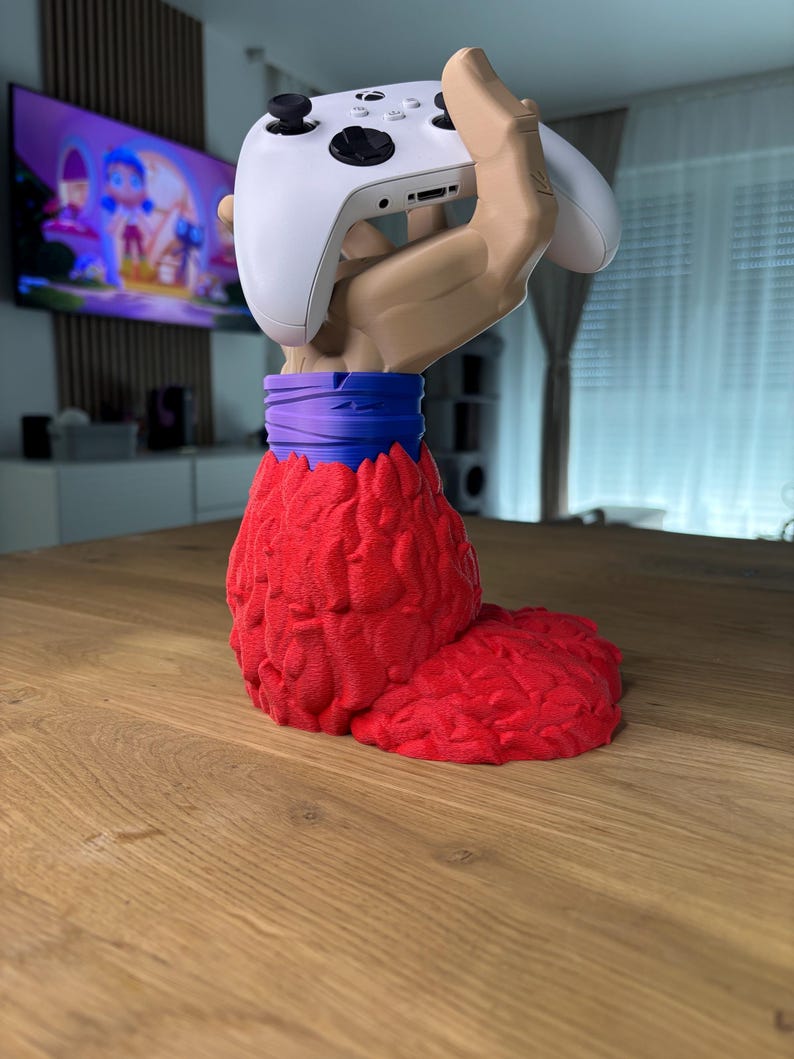 May include: A 3D printed hand holding a white video game controller. The hand is red and has a purple wristband. The hand is standing on a wooden surface.
