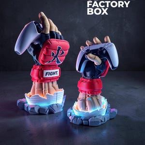 Puede incluir: Dos soportes escult&oacute;ricos con forma de manos que llevan guantes de artes marciales rojos y negros, cada uno sosteniendo un mando de videojuego blanco. Los guantes presentan el texto FIGHT y un s&iacute;mbolo kanji negro. Las bases son rocosas con luz azul.