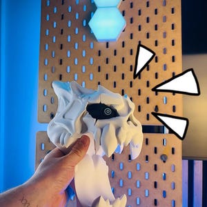 May include: A white 3D-printed skull with sharp teeth and a black eye socket is held in front of a pegboard. The pegboard is brown with evenly spaced holes. The text "3D FACTORY BOX" is visible on the bottom right.