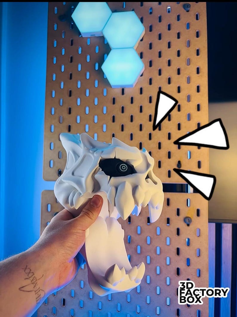 May include: A white 3D-printed skull with sharp teeth and a black eye socket is held in front of a pegboard. The pegboard is brown with evenly spaced holes. The text "3D FACTORY BOX" is visible on the bottom right.