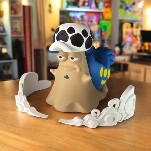 Puede incluir: Una figura de dibujos animados de color beige con un sombrero blanco y negro y una concha azul. La figura tiene una expresión de mal humor y está rodeada de accesorios blancos en forma de nube. Sobre una superficie de madera.