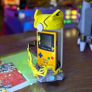 K&ouml;nnte beinhalten: Ein gelber und grauer 3D-gedruckter St&auml;nder mit Pikachu-Design h&auml;lt einen gelben Game Boy Color mit einer Pikachu-Spielkassette. Der St&auml;nder hat einen kleinen blauen Knopf auf der Vorderseite.
