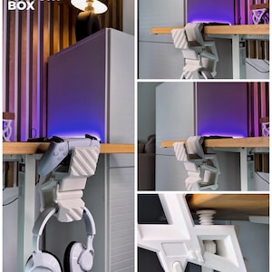 May include: A white 3D-printed desk mount for gaming accessories. The mount holds a white headset and a gray game controller. The text "3D FACTORY BOX" is visible in the upper left corner. The mount is attached to a wooden desk.