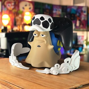 May include: A tan and blue video game controller holder shaped like a cartoon character, with a black controller resting on its back. The character wears a white hat with black spots. White decorative accents are on the sides.