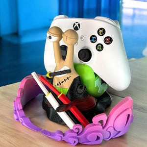 May include: A white Xbox controller rests on a custom-designed stand featuring a cartoon snail character with a sword and a pink and purple headset. The stand has a green base and holds the controller securely.