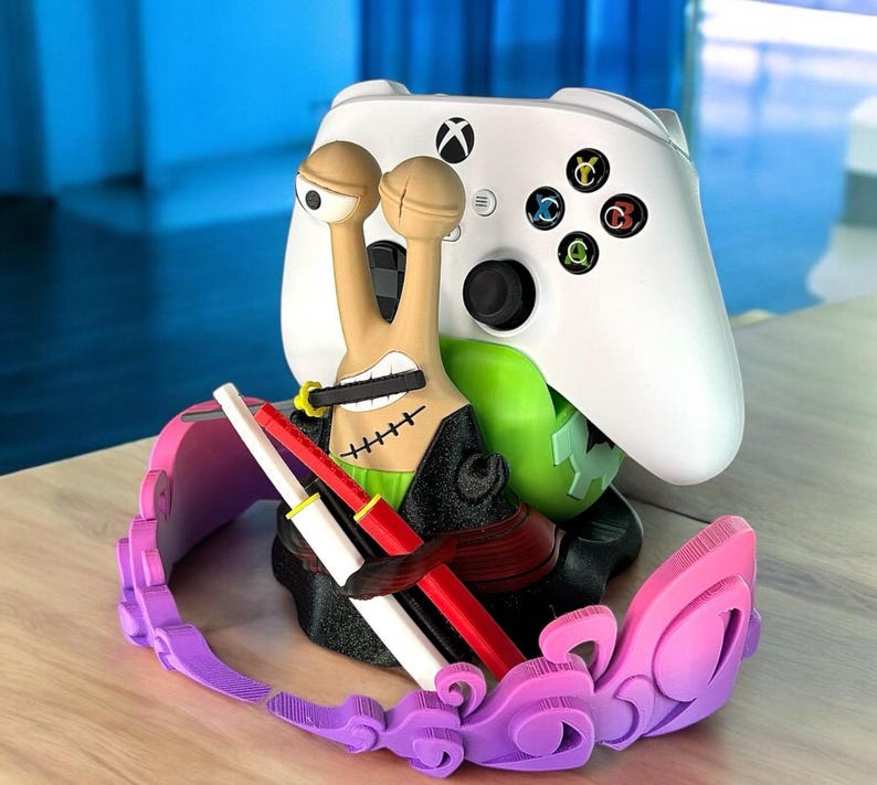 May include: A white Xbox controller rests on a custom-designed stand featuring a cartoon snail character with a sword and a pink and purple headset. The stand has a green base and holds the controller securely.