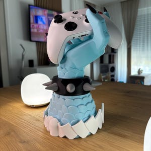 K&ouml;nnte beinhalten: Ein hellblauer, klauenf&ouml;rmiger Gaming-Controller-St&auml;nder, der einen wei&szlig;en Xbox-Controller h&auml;lt. Der St&auml;nder hat einen schwarzen Kragen mit grauen Stacheln und eine wei&szlig;e, gezackte Basis. Der Controller hat schwarze Kn&ouml;pfe und einen schwarzen Joystick.