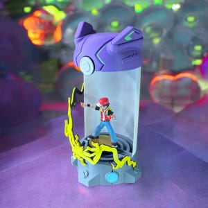 May include: A purple and gray Pokemon toy with a clear plastic tube containing a small red and black figure of a character from the Pokemon video game series. The figure is standing on a yellow platform with a lightning bolt design. The toy is on a purple surface.