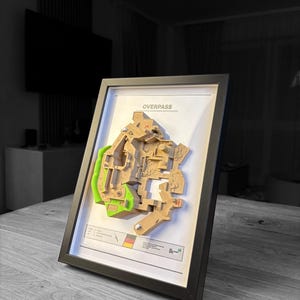 May include: Framed 3D map of a video game level, labeled "OVERPASS". The map features tan and green sections, set against a white background within a black frame. A small graphic with a German flag is visible.