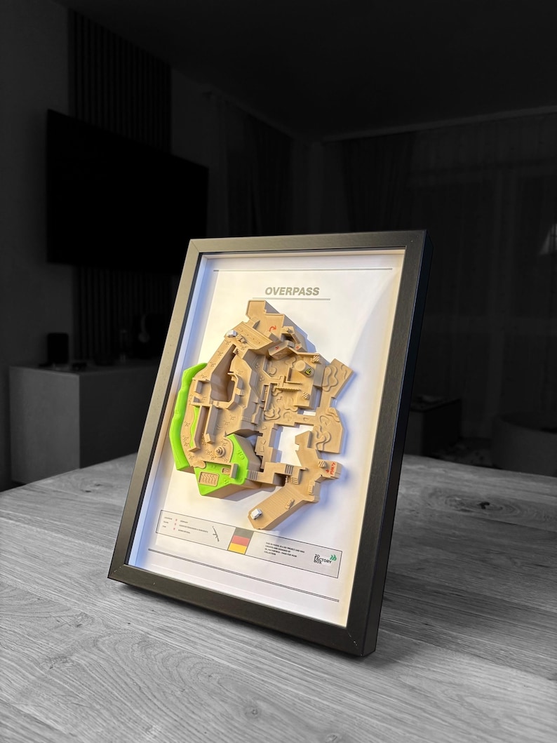 May include: Framed 3D map of a video game level, labeled "OVERPASS". The map features tan and green sections, set against a white background within a black frame. A small graphic with a German flag is visible.