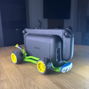 K&ouml;nnte beinhalten: Eine schwarze Nintendo Switch-Konsole, die an einem gr&uuml;n-gelben Zubeh&ouml;r im Kart-Stil befestigt ist. Der Kart hat schwarze R&auml;der mit gelben Zentren und einen blauen Sto&szlig;f&auml;nger. Die Konsole hat das Nintendo-Logo.