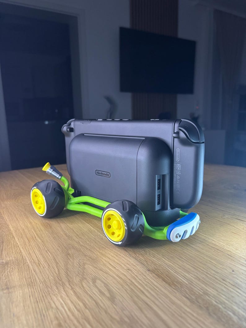 K&ouml;nnte beinhalten: Eine schwarze Nintendo Switch-Konsole, die an einem gr&uuml;n-gelben Zubeh&ouml;r im Kart-Stil befestigt ist. Der Kart hat schwarze R&auml;der mit gelben Zentren und einen blauen Sto&szlig;f&auml;nger. Die Konsole hat das Nintendo-Logo.