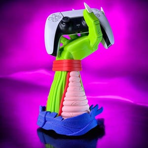 May include: A colourful gaming controller stand. The stand features a green hand holding a white and black controller. The hand is supported by a red band, a pink cone, and a blue base. The background is a gradient of purple.