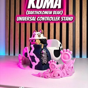 Puede incluir: Un soporte universal para controlador Kuma (Bartholomew Bear) rosa y negro. El soporte presenta un dise&ntilde;o detallado con el texto "KUMA" y "UNIVERSAL CONTROLLER STAND" en un brillo rosa ne&oacute;n.