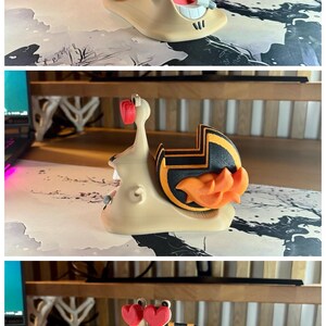 May include: A whimsical 3D-printed snail figurine with a smiling face, heart-shaped eyes, and a striped shell in black and orange. The snail has an open mouth with visible teeth and a small object in its mouth. The base is beige.