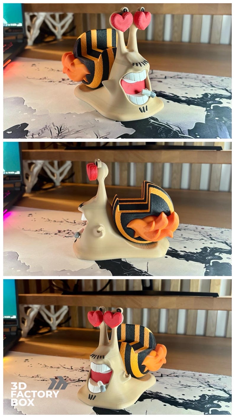 May include: A whimsical 3D-printed snail figurine with a smiling face, heart-shaped eyes, and a striped shell in black and orange. The snail has an open mouth with visible teeth and a small object in its mouth. The base is beige.