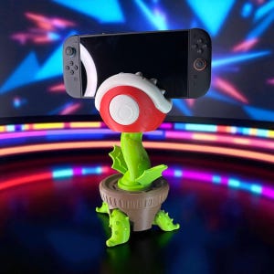 May include: A novelty plant-themed phone or gaming device stand. The stand is red, white, and green, resembling a cartoon plant. A black gaming device is held in the stand. The stand is in a brown pot with green leaves.
