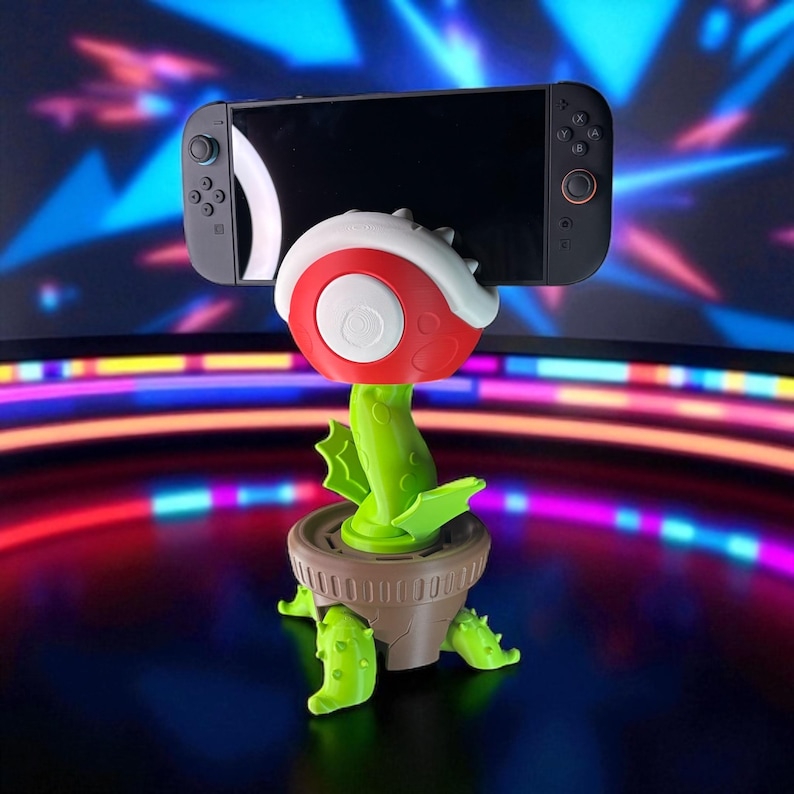 May include: A novelty plant-themed phone or gaming device stand. The stand is red, white, and green, resembling a cartoon plant. A black gaming device is held in the stand. The stand is in a brown pot with green leaves.