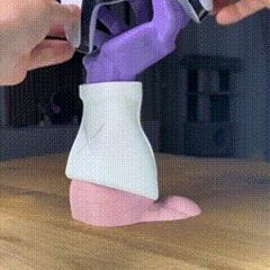 Op de afbeelding: Een wit en zwarte gamecontroller wordt vastgehouden door een paarse handvormige standaard. De standaard staat op een houten oppervlak. De standaard heeft een witte bovenkant en een roze basis. De controller is zwart en wit.