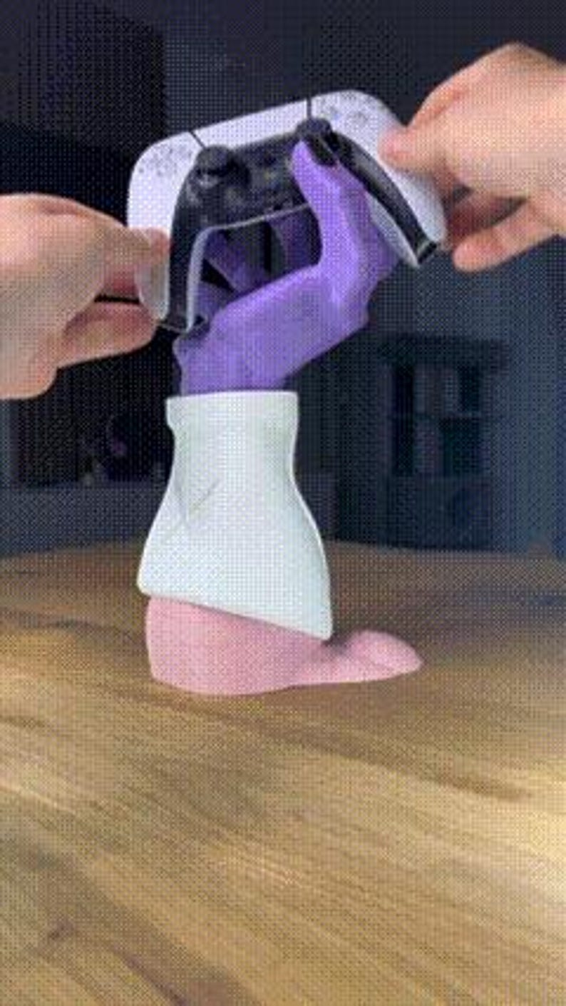 Op de afbeelding: Een wit en zwarte gamecontroller wordt vastgehouden door een paarse handvormige standaard. De standaard staat op een houten oppervlak. De standaard heeft een witte bovenkant en een roze basis. De controller is zwart en wit.