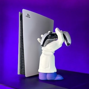 Puede incluir: Una consola PlayStation 5 blanca con una mano blanca sosteniendo un mando PlayStation 5 blanco. La mano lleva un guante azul y está de pie sobre una superficie morada.