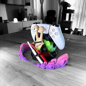 May include: A white and black video game controller is propped up by a colorful 3D-printed stand. The stand features a snail-like character with a green base, a pink and purple decorative element, and two red and white swords.