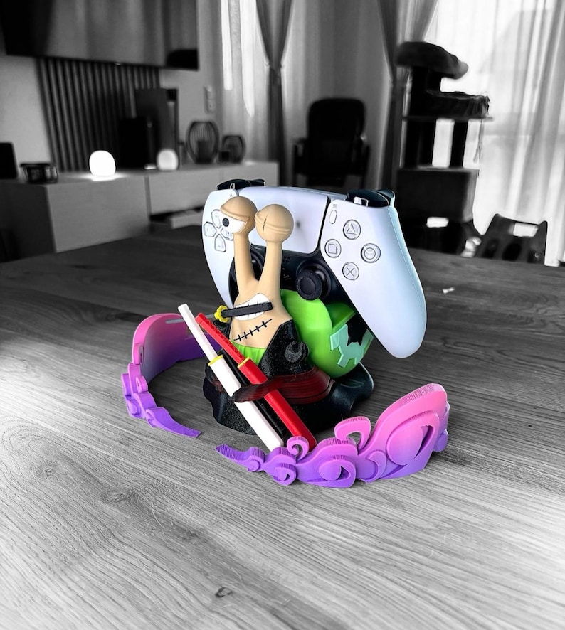 May include: A white and black video game controller is propped up by a colorful 3D-printed stand. The stand features a snail-like character with a green base, a pink and purple decorative element, and two red and white swords.
