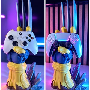 May include: A blue and yellow glove-shaped controller stand featuring three long silver claws extending upward. The base is sculpted with jagged rock-like details. The stand is shown holding a white Xbox controller and a white PlayStation controller.