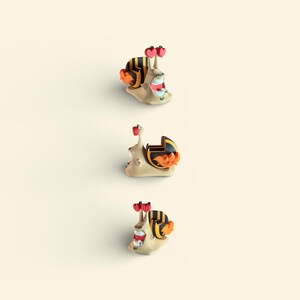 May include: Three whimsical snail figurines with beige bodies, black and yellow striped shells, and orange accents. Each snail has red heart-shaped antennae and a smiling mouth. The logo "3D FACTORY BOX" is in the lower left corner.