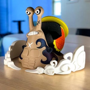 May include: A tan, cartoonish snail figurine with a stern expression, black markings, and a yellow and orange shell. The snail has large, expressive eyes and is surrounded by white cloud-like accents.