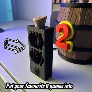 May include: A black game cartridge holder with a wooden stopper on top. The holder has slots for eight games. The text reads: "Put your favourite 8 games into the hidden games holder!" A decorative barrel and a number "2" are in the background.