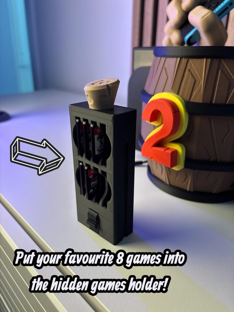 May include: A black game cartridge holder with a wooden stopper on top. The holder has slots for eight games. The text reads: "Put your favourite 8 games into the hidden games holder!" A decorative barrel and a number "2" are in the background.