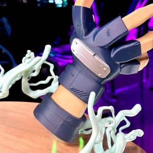 May include: A close-up of a figurine's hand, featuring a blue glove with a silver plate and tan-colored fingers. The hand is positioned above a wooden surface, with light green, abstract shapes in the foreground.