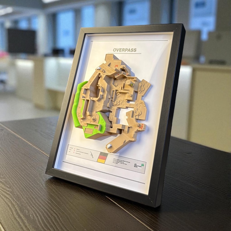 May include: Framed 3D map of the Overpass game level, featuring tan and green architectural details. The map is set against a white background within a black frame. The word "OVERPASS" is printed at the top.