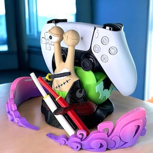 Puede incluir: Un mando blanco de PlayStation 5 descansa sobre un accesorio de juego personalizado. El accesorio presenta un diseño de caracol de dibujos animados con un atuendo negro, detalles verdes y un par de espadas. Un soporte para auriculares rosa y morado está en primer plano.