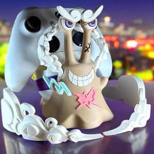 May include: A tan and white video game controller stand with a cartoon snail design. The snail has a wide grin, purple eyes, and a pink star on its body. The stand is designed to hold a white game controller.