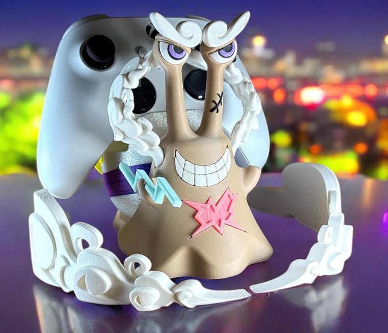 May include: A tan and white video game controller stand with a cartoon snail design. The snail has a wide grin, purple eyes, and a pink star on its body. The stand is designed to hold a white game controller.