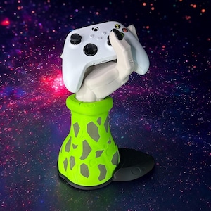 May include: A green and grey hand-shaped controller stand with a white Xbox controller in the hand. The stand is on a black base.