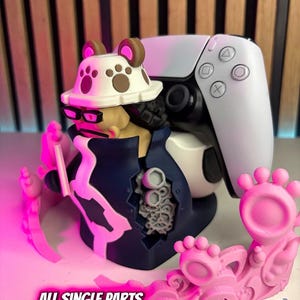 Puede incluir: Un mando de PlayStation 5 blanco y negro se combina con una figura impresa en 3D de un personaje que lleva sombrero y gafas. La figura es azul, amarilla y blanca. Elementos decorativos rosas rodean la base. El texto de la imagen dice: "ALL SINGLE PARTS ARE FITTING PERFECT!"