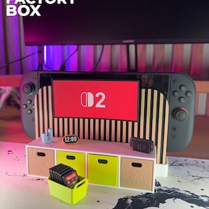 May include: A 3D Factory Box display featuring a Nintendo Switch console with a red screen displaying the "D2" logo. The display includes a small white cabinet with lime green and tan drawers, and a lime green container holding game cartridges. The time 12:00 is displayed.