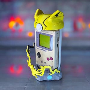 K&ouml;nnte beinhalten: Eine gelbe und graue Game Boy Color Konsole mit einer gelben und grauen Pikachu Figur auf der Oberseite. Die Konsole steht auf einem grauen St&auml;nder mit einer gelben und grauen Pikachu Figur an der Seite.